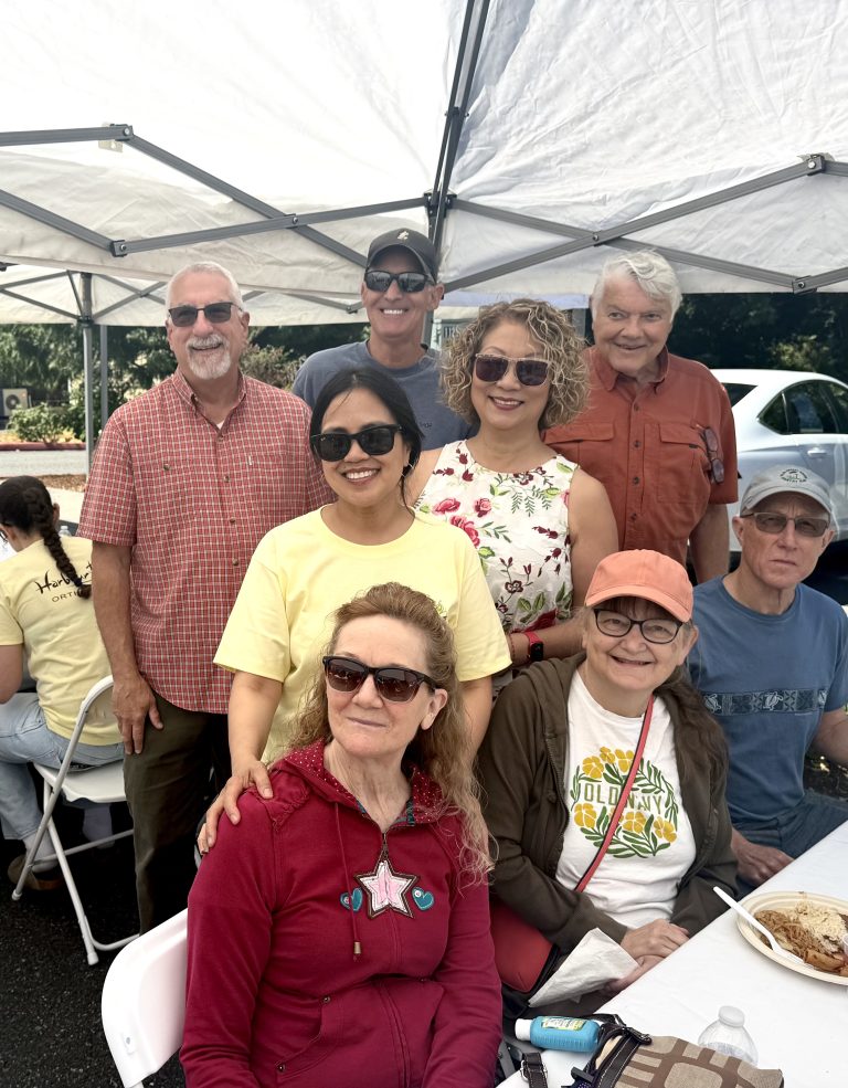 Group of smiling adults posing under a white canopy at an outdoor event, wearing casual clothes and sunglasses.