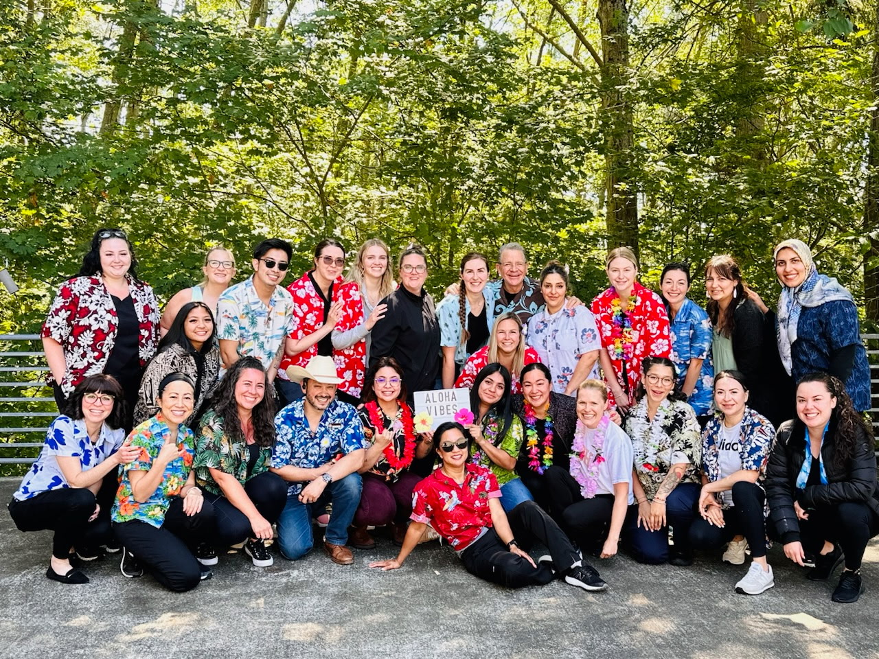Group of diverse people in colorful Hawaiian shirts posing together outdoors, a sign reads 'Aloha Vibes' at center.
